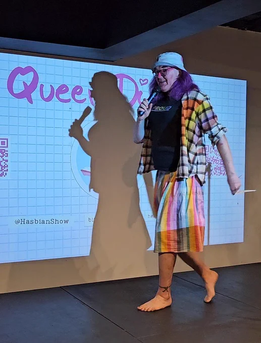 A person with purple hair, a plaid overshirt, black t-shirt, striped rainbow skirt, and a blue bucket hat is speaking barefoot on stage. They hold a microphone, AI tells me in a way that's "projecting confidence". YMMV.  The phrase "Queer Diary" is on a projection screen in pink letters behind them.