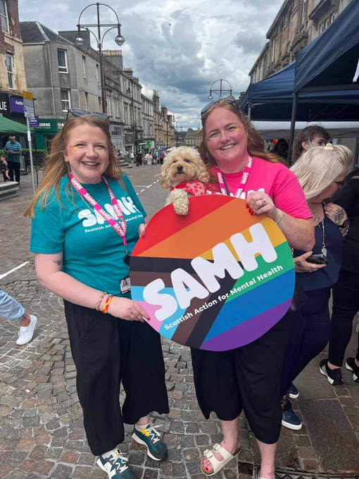 Catherine and Heather (and a cute doggo) hold a SAMH logo in LGBTQI  colours