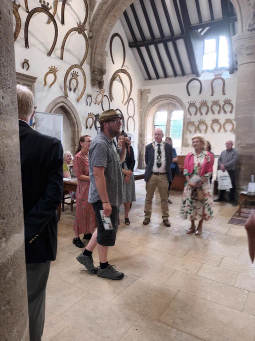 An old building with brown beams and large horse shoes in a white wall.  A group of people are standing in a semi circle facing to the right