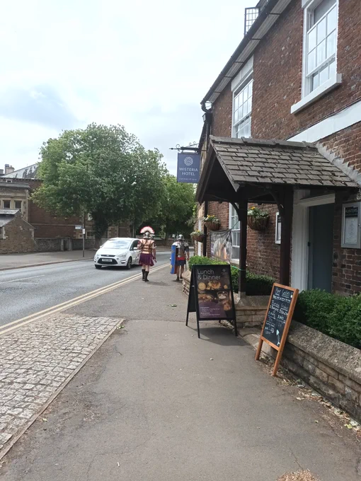 2 people dressed up as Romans walking past a red brick building with a porch, they are on a pavement next to a road