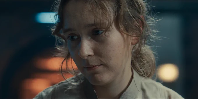 A close-up of Aliss, a young woman with a dirty face, tousled hair and a thoughtful, slightly downcast expression. 
