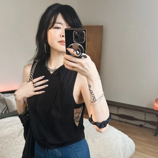 Ruibo Qian stands in a black blouse and blue slacks, holding up a phone to a mirror. 