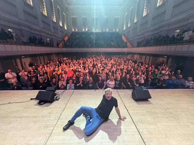 Rhys darby poses on stage in front of a large crowd in the UK