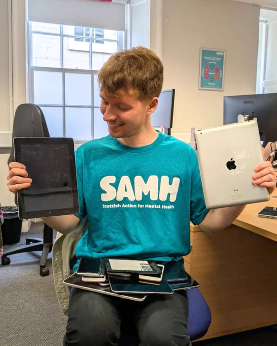 A smiling SAMH staff member proudly holds up a collection of old tech, including two iPads. He's wearing a teal SAMH tshirt.
