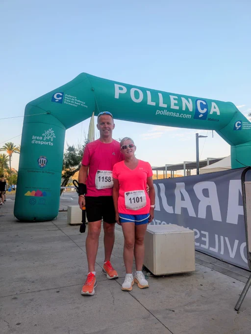 Kelly and Mike stood in front of the start line both wearing running clothes and race numbers - the sky behind them is blue