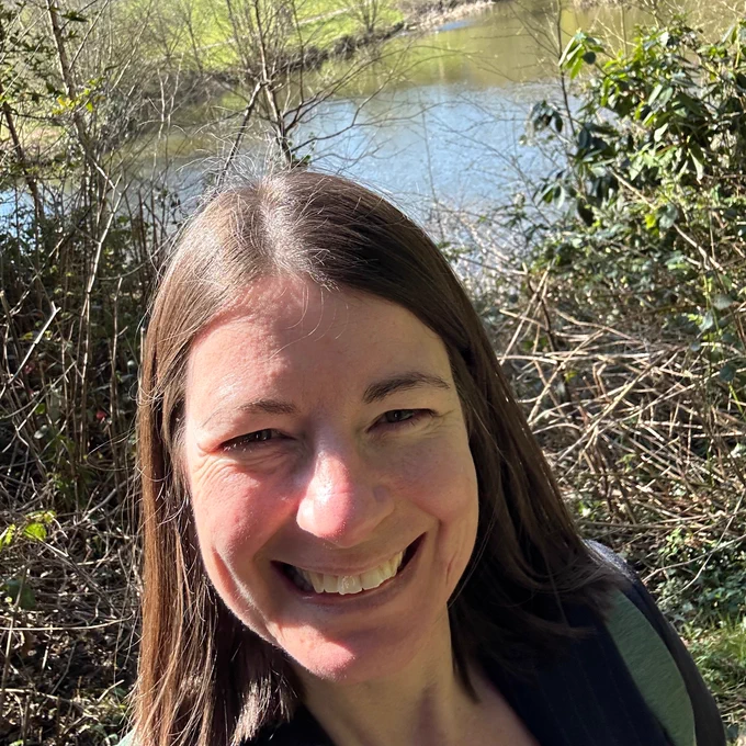 smiling middle aged brown haired woman, standing in front of a lake surrounded by bushes