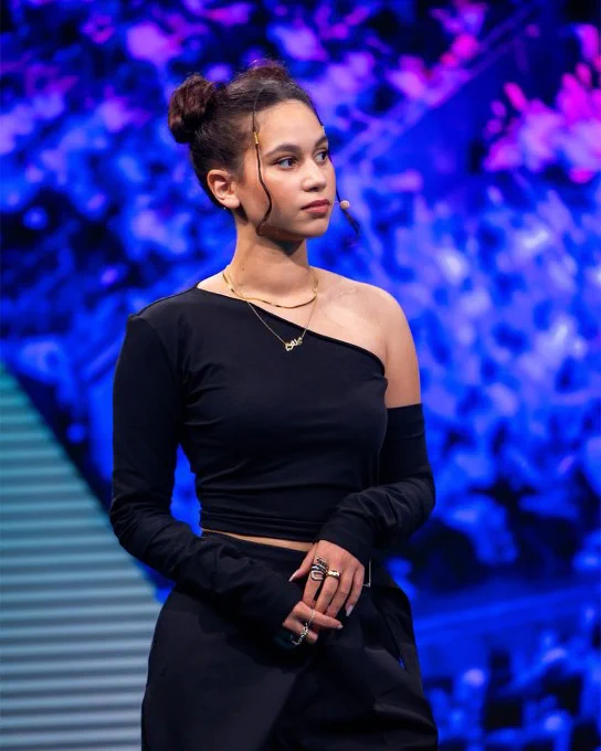 Keltoum Baddaje wearing a stylish black one-shoulder top and multiple rings, standing against a vibrant blue and purple abstract background.