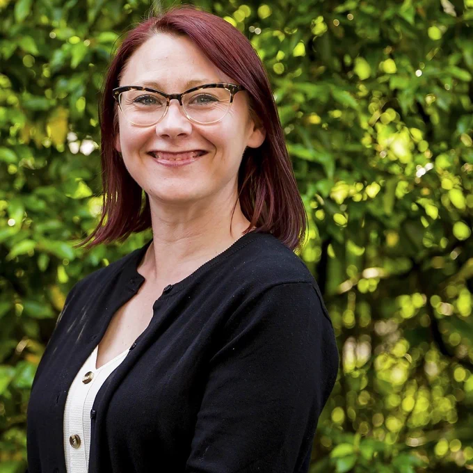 Zoe Davison, Director of Research and Information at BCRT,  wearing a black cardigan over a white buttoned top standing outdoors against green foliage.