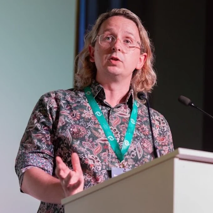 Ashley Ball-Gamble, CEO at CCLG, wearing a floral patterned shirt and green lanyard speaking at a podium with a microphone.