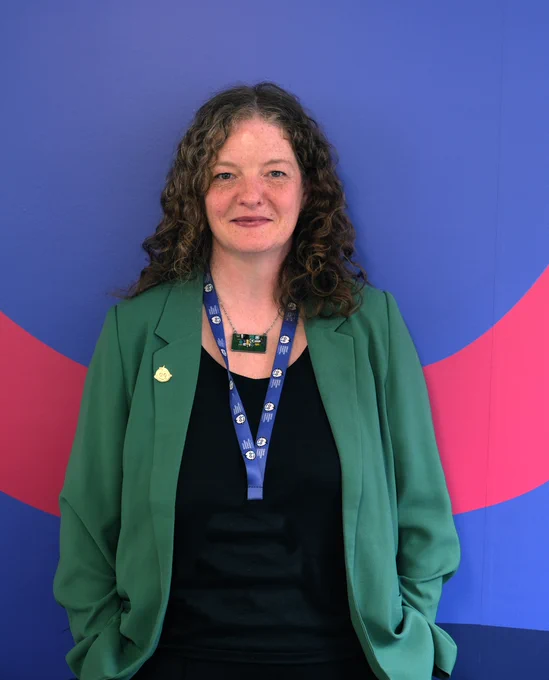 Aoife Regan, Director of Impact and Charitable Programmes at Great Ormond Street Hospital Charity, wearing a green blazer over a black top with a blue lanyard against a colorful blue and pink background.