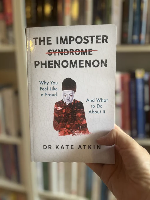 Book, titled The Imposter Phenomenon (with the word syndrome struck out) held in a hand with bookshelves in the background. Subtitle is 'why you feel like a fraud and what to do about it'. Cover also features a figure in red holding a mask in front of their face. By Dr Kate Atkin
