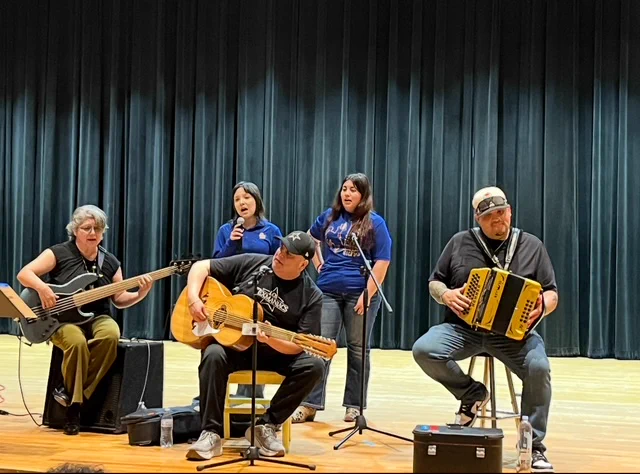 Three seated adults play guitar, bajo sexto, and accordion while two students stand behind them and sing. They are on a stage, with a grey-blue velvet curtain behind them.