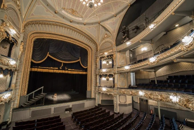 Looking towards the ornate stage the Tyne Theatre & Opera House with gilded decorative details, a grand proscenium arch framing a stage with dark blue curtains, tiered balconies with plush seating, and an elegant domed ceiling featuring intricate plasterwork and a central chandelier.