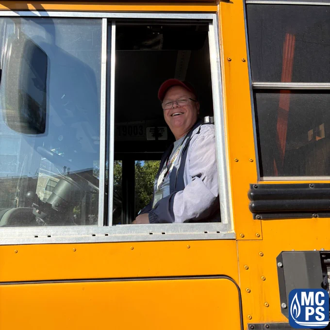 School bus driver wearing cap and light jacket sitting inside the yellow bus near the window.