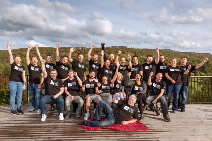 A large group of enthusiastic people wearing matching “CodeCabin 2018” black t-shirts pose outdoors on a wooden deck. Many have their arms raised in celebration. The group is surrounded by lush green hills under a partly cloudy sky, with one person playfully lying on a red picnic blanket in front.
