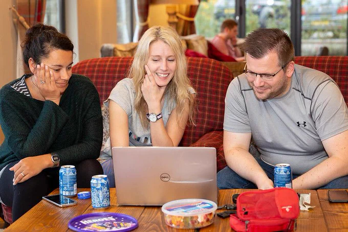 Three people sit on a cozy red plaid couch, smiling and focused on a Dell laptop. Two women and one man lean in, enjoying a relaxed collaboration. The table in front of them holds soda cans, snacks, and a red pouch. A casual, homey atmosphere is visible, with others chatting in the background.