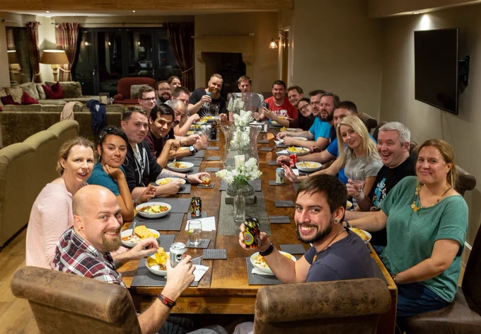 A large group of people sits around a long wooden dining table, smiling and enjoying a shared meal in a cozy indoor setting. Plates of food, drinks, and decorative flowers fill the table, and many attendees raise glasses or cans in a toast. The relaxed atmosphere suggests a retreat or community event.