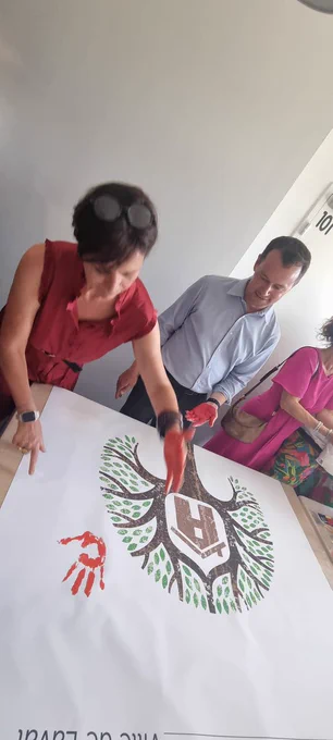 Madame la préfète et une autre personne apposent l'empreinte de leurs mains peintes en rouge sur une feuille posée devant eux sur laquelle figure le dessin d'une maison dans un arbre symbolisant la maison des 1000 premiers jours.