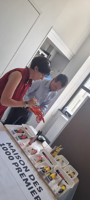 Madame la préfète et une autre personne peignent leur main en rouge pour laisser une empreinte sur une feuille posée devant eux.