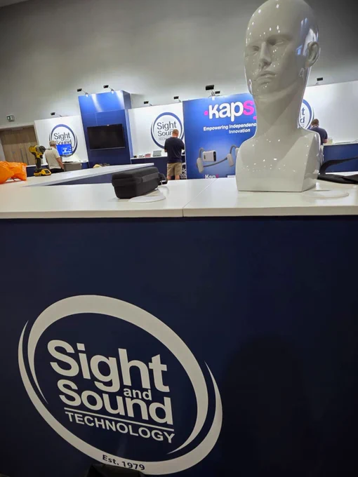 A view of the Sight and Sound Technology booth at an exhibition. In the foreground is a sleek, glossy white mannequin head used for display, positioned on a blue counter branded with the company’s logo and tagline “Sight and Sound Technology – Est. 1979.” In the background, people are setting up other displays including the Kapsys booth and screens. The environment is professional and well-lit.