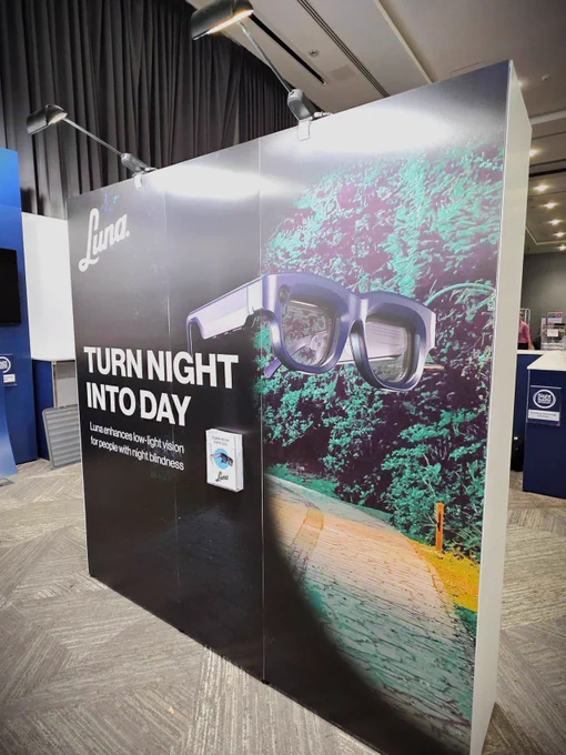 A promotional display for Luna, a device designed to enhance low-light vision for people with night blindness. The banner reads “TURN NIGHT INTO DAY,” with the product image showing a pair of high-tech glasses against a dark forest pathway illuminated in false color to simulate enhanced visibility. Overhead spotlights highlight the bold graphics and product messaging. 