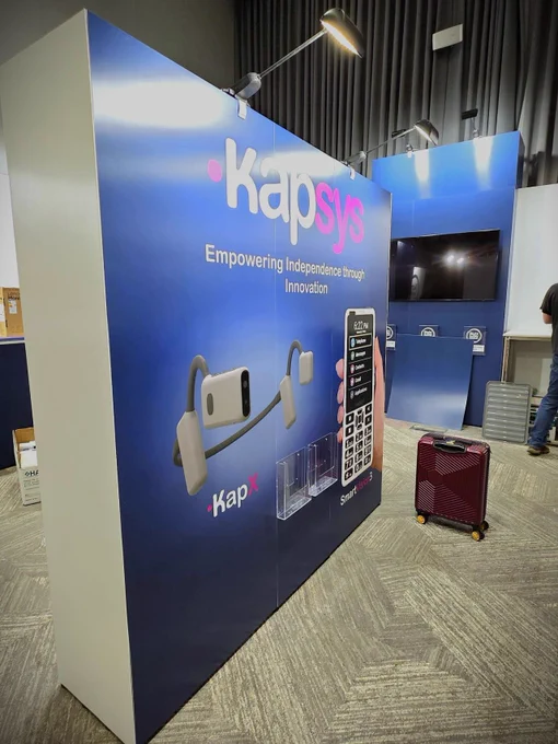 A trade show booth for Kapsys, featuring a dark blue backdrop with the brand's logo in white and pink. The slogan reads “Empowering Independence through Innovation.” A product called KapX is prominently displayed, appearing to be a wearable device. A hand is holding a smartphone. There are clear acrylic holders attached to the display wall. 