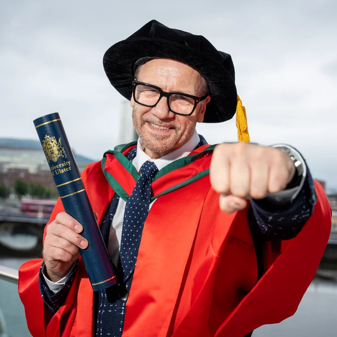 Honorary Graduate Billy Murray in a red academic gown and black hat holding an University of Ulster diploma case by a riverside cityscape.