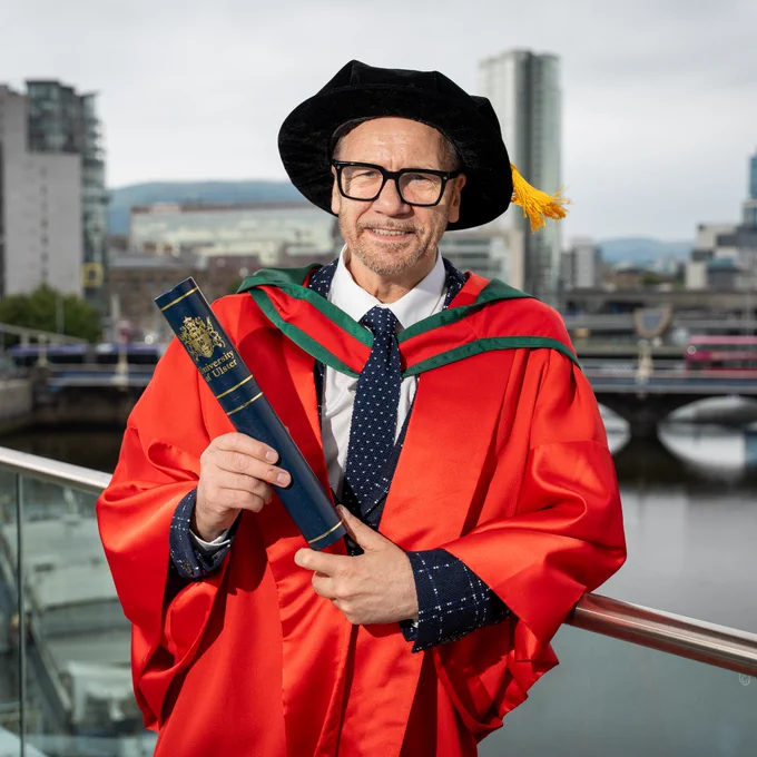 Honorary Graduate Billy Murray in a red academic gown and black hat holding an University of Ulster diploma case by a riverside cityscape.