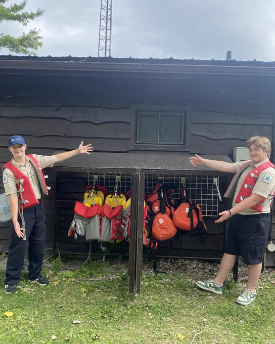 Two park staff wearing lifejackets, smiling and standing with their thumbs up in front of a rack of lifejackets in front of a brown office building.