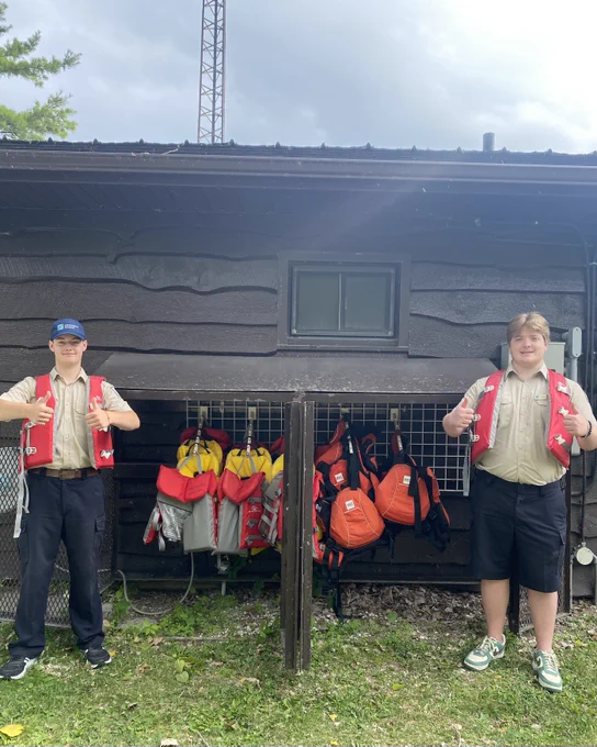 Two park staff wearing lifejackets, smiling and gesturing with their hands towards a rack of lifejackets in front of a brown office building.