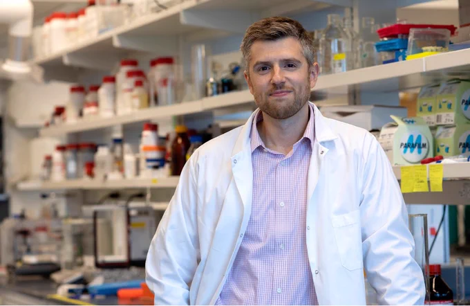 Daniel Roxbury wearing a white lab coat as he stands in a laboratory filled with chemical bottles and scientific equipment on shelves behind him.