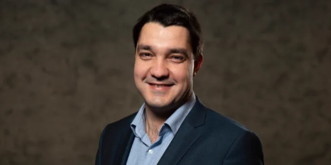 Portrait of Radoslav Ivanov smiling, wearing a blue blazer over a blue shirt, set against a textured gray backdrop.