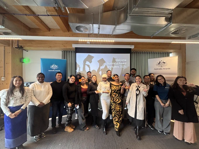 18 scholars all smiling and dressed in semi-formal clothes standing inside a room in front of two Australia Awards banners and screen saying: Welcome Australia Awards graduates 2025! 