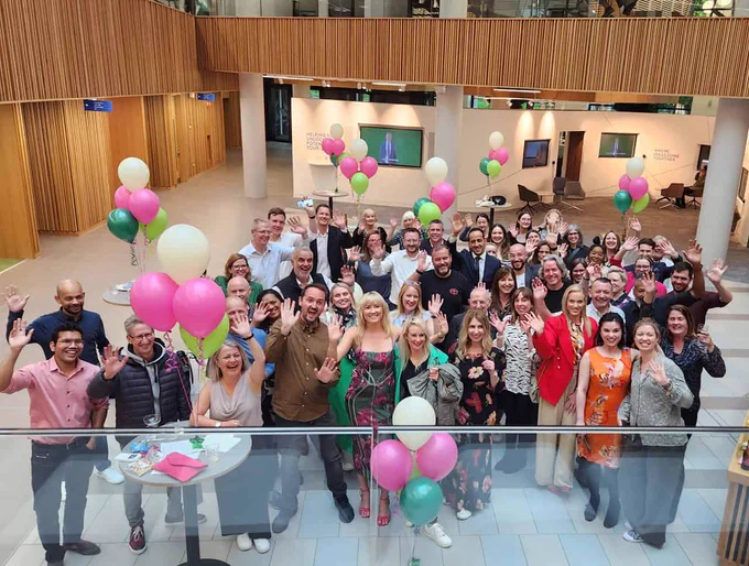 Image is a photo of a large group of people having a celebration in an indoor large space.  Some are holding balloons.