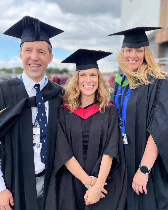 Three graduates wearing black caps and gowns stand together outdoors under a cloudy sky during a graduation ceremony.