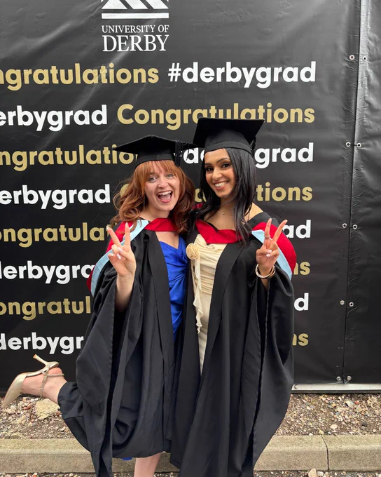 Two graduates stand together doing peace signs and smiling at the camera. Behind them is a banner which says 'Congratulations #DerbyGrad'