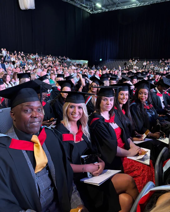 Graduates in black gowns seated in rows at a large indoor ceremony with an audience in the background. A row of graduates smile and look at the camera.