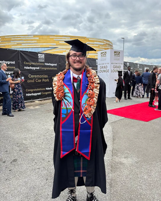 Graduate in black gown and cap stands on pavement outside University of Derby's Grad Lounge with a coulorful stole and floral lei.