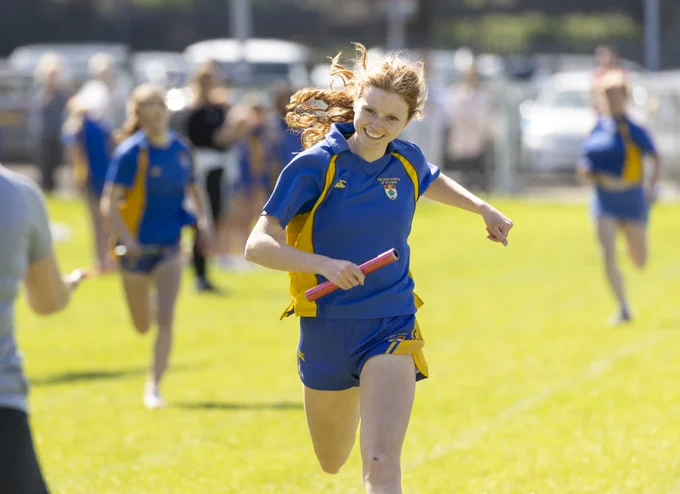 A female HSOG pupil athlete in a blue and yellow sports uniform runs across a grass field while holding a red relay baton, her hair flowing behind her as she smiles. She is participating in a track and field relay race, with other pupils in matching blue uniforms visible in the blurred background.