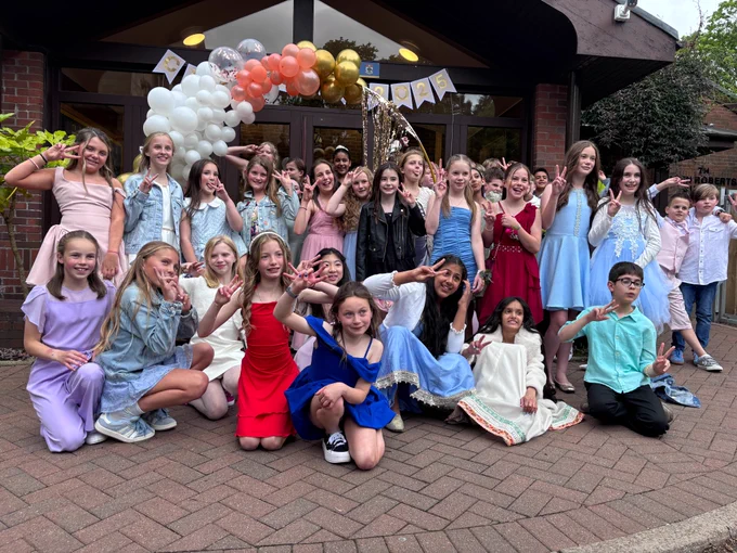 A large group of J6 pupils in formal attire posing for a group photo outside the Junior School, with decorative balloons. The pupilsare wearing colourful dresses and formal clothing in shades of blue, pink, red, white and purple. Many are making peace signs with their hands and smiling at the camera. Behind them is a balloon arch made of white, coral and gold balloons, along with bunting flags.