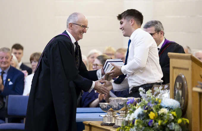 Rector, John O'Neill, in black academic robes with purple trim shakes hands with a male pupil in a white shirt and blue tie while presenting him with books. The ceremony takes place on a stage with decorative flower arrangements, silver trophies and a wooden podium visible. An audience of staff can be seen seated in the background, with Mr Niall Campbell partially visible on the right side of the image.