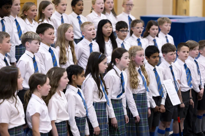 A large group of HSOG Junior School pupils in uniform performing as a choir, arranged in tiered rows. The pupils are wearing white shirts with blue and yellow striped ties and dark tartan skirts or trousers. They are singing with their mouths open, engaged in their performance.