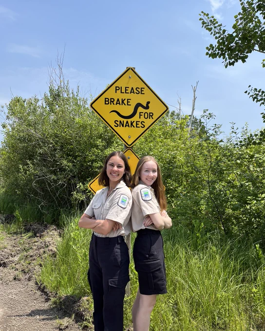 Two park staff smiling and standing back-to-back beneath a bright yellow road sign that reads: "PLEASE BRAKE FOR SNAKES".