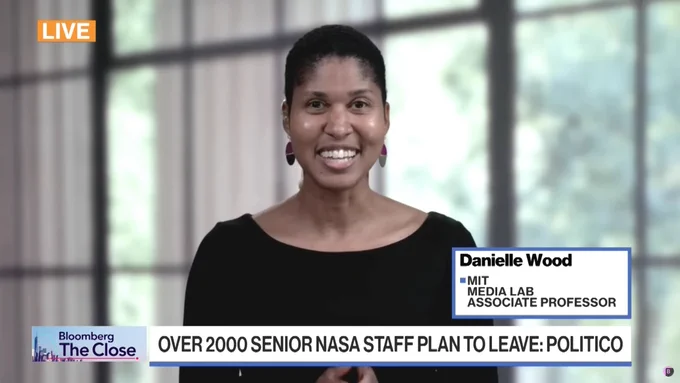 Screenshot from Bloomberg interview with Media Lab Professor Danielle Wood. Professor Wood sits in front of a large window, through which blurred columns and trees are visible. Professor Wood is in sharp focus, smiling and wearing a black shirt. A card in the lower-right corner identifies her as "Danielle Wood, MIT Media Lab Associate Professor." A chyron at the bottom of the screen has the title of the show, "Bloomberg: The Close," on the left, and the words "Over 2000 Senior NASA staff plan to leave: Politico" on the right. 