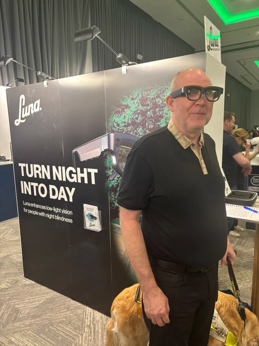 A man with a guide dog stands in front of a booth for Luna, a company offering low-light vision glasses. The booth display reads “TURN NIGHT INTO DAY,” highlighting the purpose of the glasses.