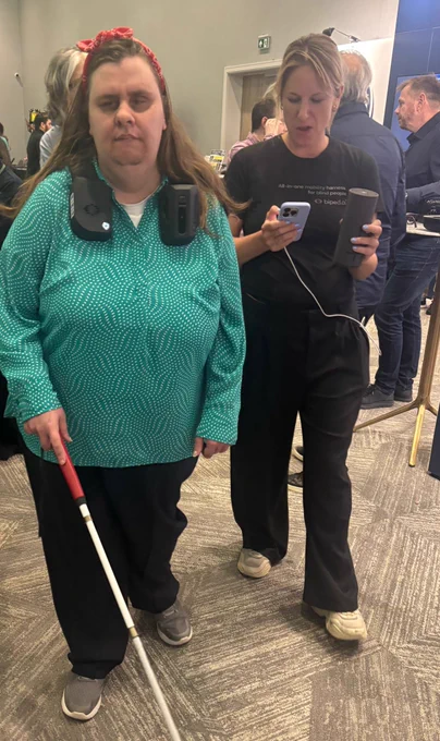 A woman with a white cane is accompanied by another woman holding a phone and a cylindrical device. The blind woman wears a green polka-dot blouse and has a wearable assistive device around her neck.