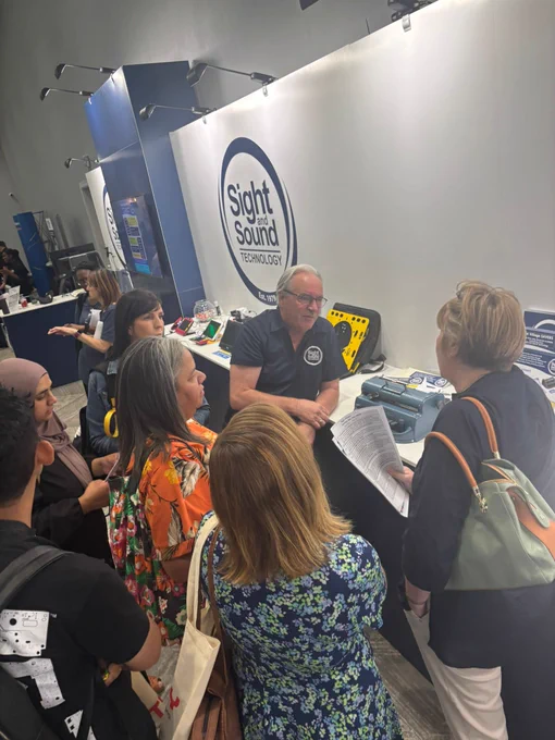 A large group gathers around a booth hosted by Sight and Sound Technology. CEO Glenn Tookey explains assistive tools, including a Perkins brailler. The group includes women in colorful attire and a man in a branded polo.