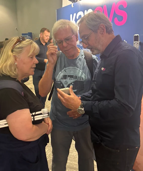 Three people are examining a KAPSYS phone together. The man in a dark shirt, representing the company "Kapsys," appears to be explaining an accessible mobile device to the couple.