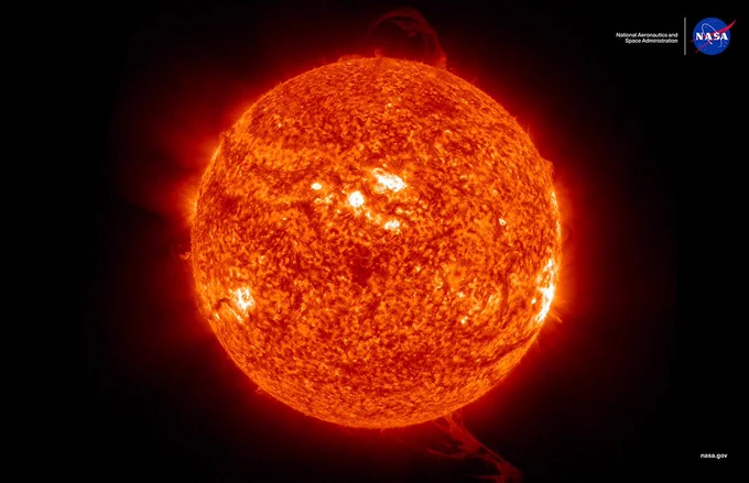 A detailed image of the Sun glowing bright orange-red with visible solar flares and active regions; NASA's logo appears in the top right corner on a black background.