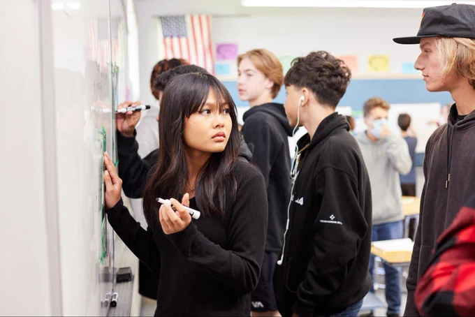 Students in a classroom facing and writing on a whiteboard on the wall.